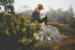 Woman in flannel baseball hat on rock writing in journal